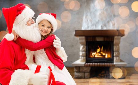 holidays, christmas, winter and people concept - happy girl with santa claus and gift box over home fireplace and lights backgroundの写真素材