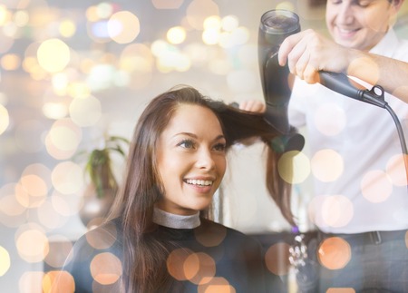 beauty, hairstyle and people concept - happy young woman and hairdresser with fan making hot styling at hair salon over holidays lightsの写真素材