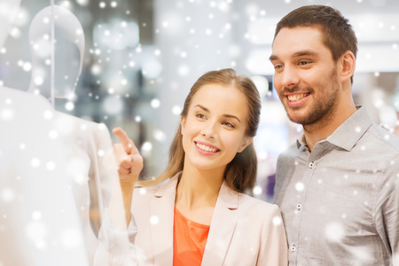 sale, consumerism and people concept - happy young couple pointing finger to shop window in mall with snow effectの写真素材
