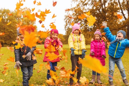 childhood, leisure, friendship and people concept - group of happy kids playing with autumn maple leaves and having fun in park