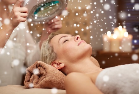 people, beauty, skin care, winter and relaxation concept - close up of beautiful young woman lying with closed eyes and beautician looking through magnifying lamp in spa salon with snow effectの写真素材