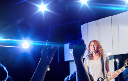 nightlife, music, technology and people concept - close up of singer playing electric guitar and singing on stage over fans crowd taking picture or video by tablet pc computer at concert in night clubの写真素材