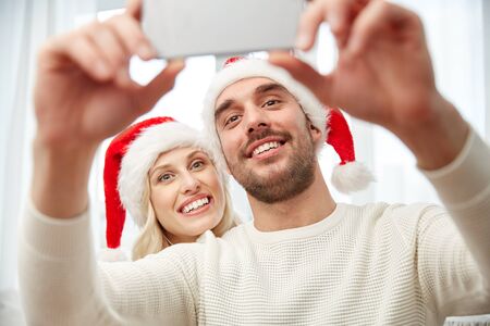 christmas, holidays, technology and people concept - happy couple in santa hats taking selfie picture with smartphone at homeの写真素材