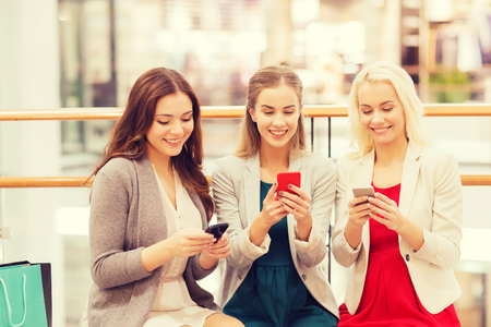sale, consumerism, technology and people concept - happy young women with smartphones and shopping bags in mallの写真素材