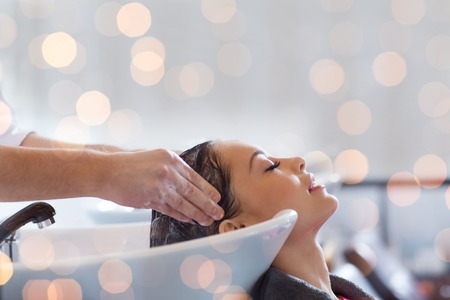 beauty, hair care and people concept - happy young woman with hairdresser washing head at hair salon over holidays lightsの写真素材