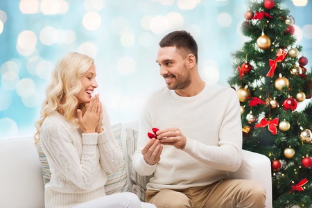 love, christmas, couple, proposal and people concept - happy man giving engagement ring in little red box to woman over blue holidays lights backgroundの写真素材