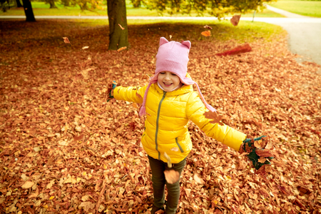 autumn, childhood, nature and people concept - happy little girl playing with fallen leaves in parkの写真素材