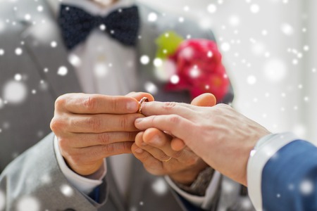 people, homosexuality, same-sex marriage and love concept - close up of happy male gay couple hands putting wedding ring on over snow effectの写真素材