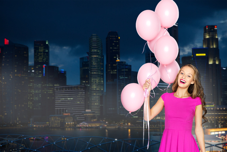 people, holidays, party, nightlife and fashion concept - happy young woman or teen girl in pink dress with helium air balloons over night singapore city backgroundの写真素材