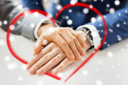 people, homosexuality, same-sex marriage and love concept - close up of happy male gay couple hands with wedding rings on and red heart shape over snow effectの写真素材