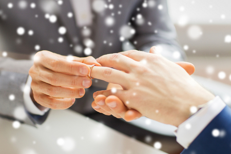 people, homosexuality, same-sex marriage and love concept - close up of happy male gay couple hands putting wedding ring on over snow effectの写真素材