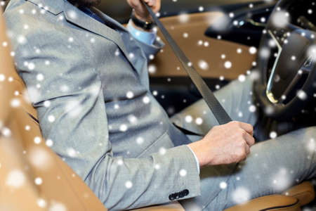 safety, driving and people concept - close up of man in elegant business suit fastening seat safety belt in car over snow effectの写真素材