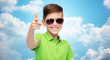 childhood, fashion, accessory, style and people concept - happy smiling boy in sunglasses and green polo t-shirt over blue sky and clouds backgroundの写真素材