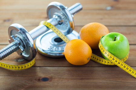sport, fitness, diet and objects concept - close up of dumbbell and green apple with oranges wrapped by measuring tape on wooden tableの写真素材