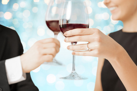 people, holidays, marriage and celebration concept - happy engaged couple clinking wine glasses over blue holidays lights backgroundの写真素材