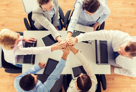 business, people, technology, cooperation and team work concept - close up of creative team with laptop and tablet pc computers sitting at table and holding hands on top of each other in officeの写真素材