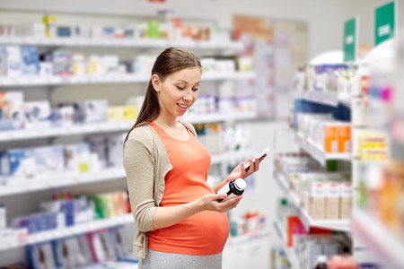 pregnancy, medicine, pharmaceutics, health care and people concept - happy pregnant woman with smartphone choosing medication at pharmacyの写真素材