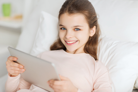 people, children, rest and technology concept - happy smiling girl lying awake with tablet pc computer in bed at homeの写真素材