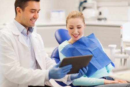 people, medicine, stomatology and health care concept - happy male dentist showing tablet pc computer to woman patient at dental clinic officeの写真素材