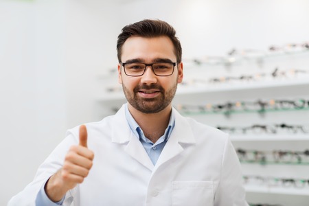health care, people, eyesight and vision concept - smiling man optician in glasses and white coat showing thumbs up gesture at optics storeの写真素材