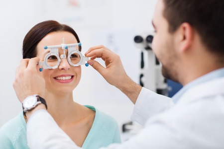 health care, medicine, people, eyesight and technology concept - optometrist with trial frame checking patient vision at eye clinic or optics store