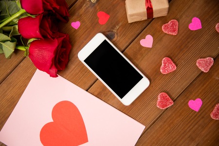 romance, valentines day and holidays concept - close up of smartphone, gift box, red roses and greeting card with heart-shaped candies on woodの写真素材