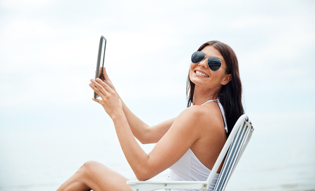 summer vacation, tourism, travel, holidays and people concept - smiling young woman with tablet pc computer sunbathing in lounge or folding chair on beachの写真素材