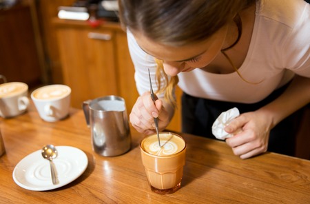 equipment, coffee shop, people and technology concept - close up of woman pouring cream to cup of coffee at cafe bar or restaurant kitchenの写真素材