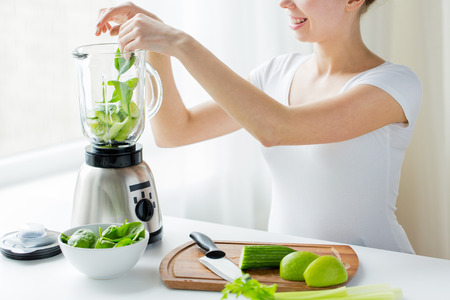healthy eating, cooking, vegetarian food, dieting and people concept - close up of young woman with blender and green vegetables making detox shake or smoothie at homeの写真素材