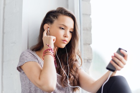 people, technology and teens concept - sad pretty teenage girl sitting on windowsill with smartphone and earphones listening to musicの写真素材