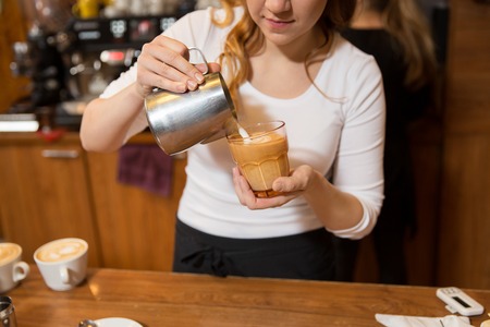equipment, coffee shop, people and technology concept - close up of woman pouring cream to cup of coffee at cafe bar or restaurant kitchenの写真素材