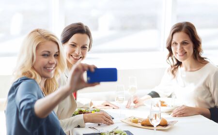 people, holidays, celebration and lifestyle concept - happy women with smartphone taking selfie at restaurantの写真素材