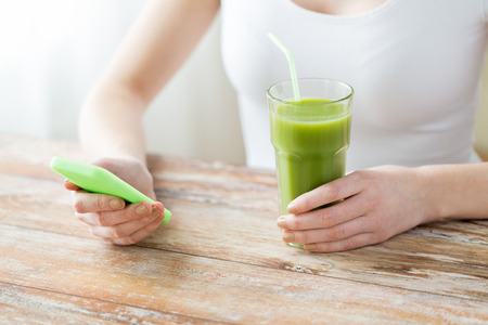 healthy eating, diet, detox, technology and people concept - close up of woman with smartphone green juice sitting at wooden tableの写真素材