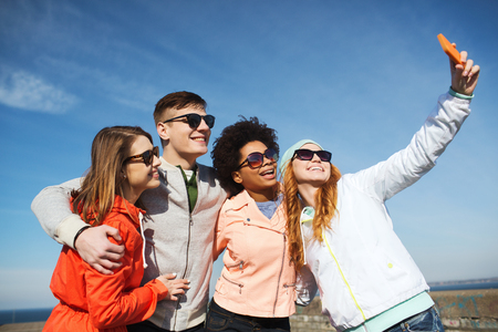 people, leisure, friendship and technology concept - group of smiling teenage friends taking selfie with smartphone outdoorsの写真素材