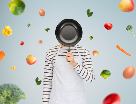 people, cooking, culinary and identity concept - man or cook in apron hiding his face behind frying pan over gray background with falling vegetablesの写真素材