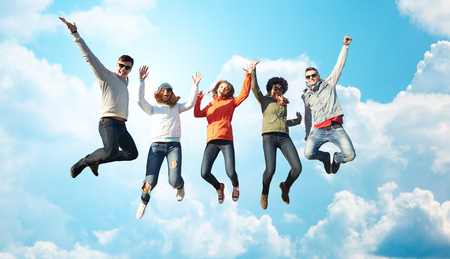 people, freedom, happiness and teenage concept - group of happy friends in sunglasses jumping high over blue sky and clouds backgroundの写真素材