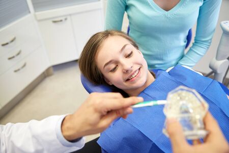 people, medicine, stomatology and health care concept - happy male dentist with toothbrush and jaw layout showing how to brush teeth to patient girl and her mother at dental clinic officeの写真素材