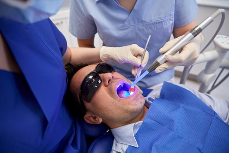 people, medicine, stomatology and health care concept - female dentist and assistant with dental curing light and mirror treating male patient teeth at dental clinic officeの写真素材