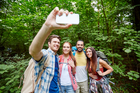 technology, travel, tourism, hike and people concept - group of smiling friends walking with backpacks taking selfie by smartphone in woodsの写真素材