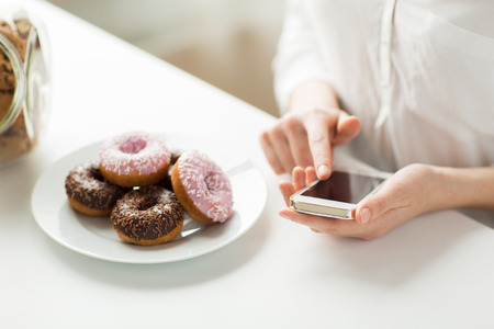 people, junk food, diet, technology and unhealthy eating concept - close up of hands with smart phone and donuts counting calories and sitting at table at homeの写真素材