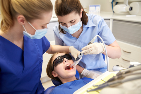 people, medicine, stomatology and health care concept - female dentists with mirror and suction treating patient girl teeth at dental clinic officeの写真素材