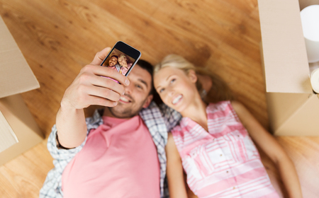 new home, technology, people, repair and moving concept - happy couple taking selfie with smartphone and lying on floor among cardboard boxes at homeの写真素材
