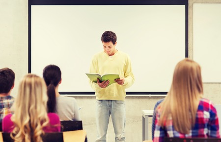 education, high school, teamwork and people concept - smiling student boy with notebook standing and reading in front of students in classroomの写真素材
