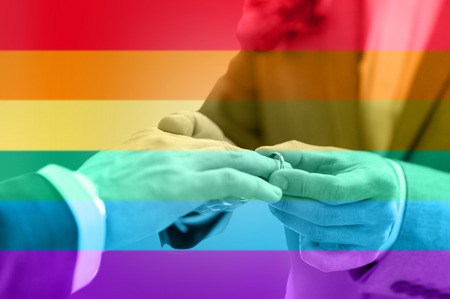 people, homosexuality, same-sex marriage and love concept - close up of happy male gay couple hands putting wedding ring on over rainbow flag stripes backgroundの写真素材