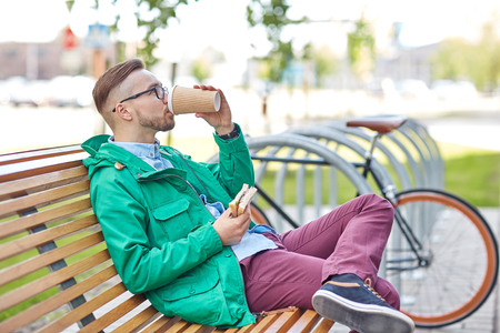 people, style, leisure and lifestyle - happy young hipster man drinking coffee cup and eating sandwich on city streetの写真素材