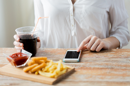 fast food, people, technology and diet concept - close up of woman with smartphone drinking coca cola and eating french fries, ketchup and deep-fried squid rings at wooden table