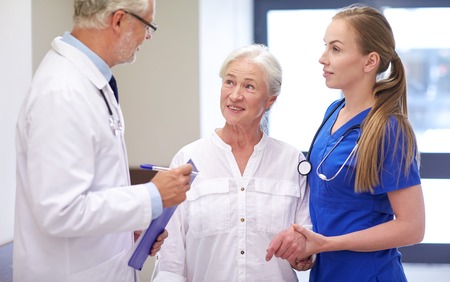 medicine, age, health care and people concept - male doctor with clipboard, young nurse and senior woman patient talking at hospital corridorの写真素材