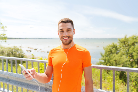 fitness, sport, people, technology and healthy lifestyle concept - smiling young man with smartphone and earphones listening to music at summer seasideの写真素材