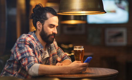 people and technology concept - happy man with smartphone drinking beer and reading message at bar or pubの写真素材