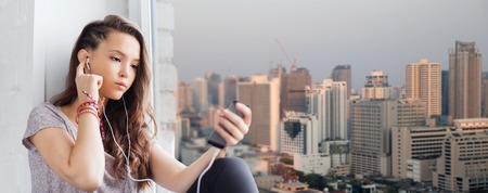 people, technology and teens concept - sad pretty teenage girl sitting on windowsill with smartphone and earphones listening to music over city backgroundの写真素材